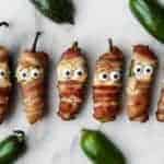 Six bacon wrapped jalapeno poppers, decorated with candy eyes to resemble mummies, are arranged on a white surface and surrounded by whole jalapeño peppers.