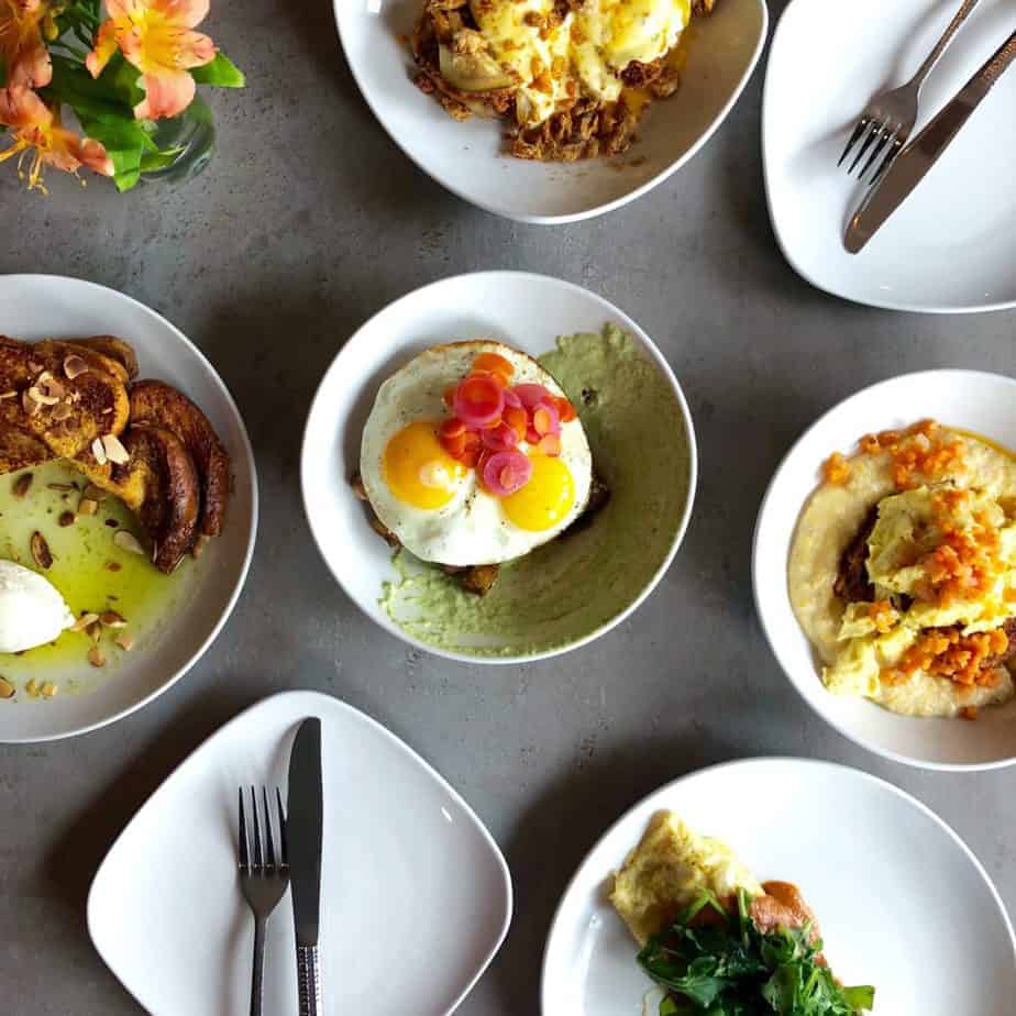A table with five plates of assorted breakfast dishes, including eggs, toast, and vegetables, is arranged on a gray surface with a flower vase in the corner—perfect for experiencing Charlotte's best brunch. Utensils rest atop each plate.