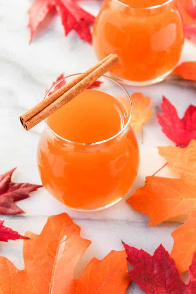 glass of spiked apple cider orange liquid topped with a cinnamon stick on a marble background with red and orange leaves