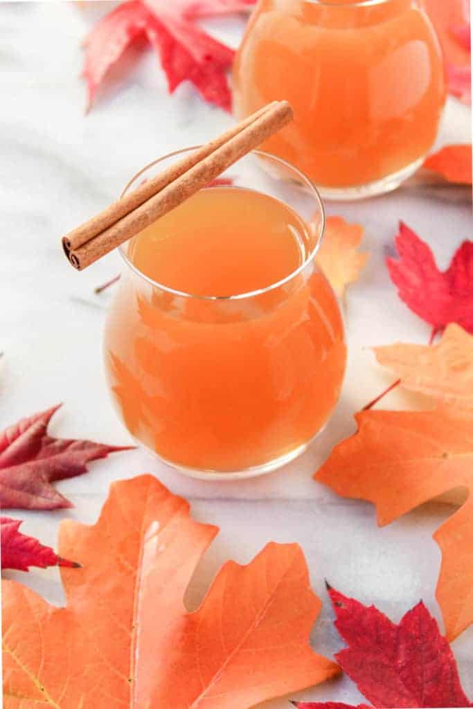 two glasses of whiskey apple cider cocktails with cinnamon sticks on a background of vibrant red and orange fall leaves