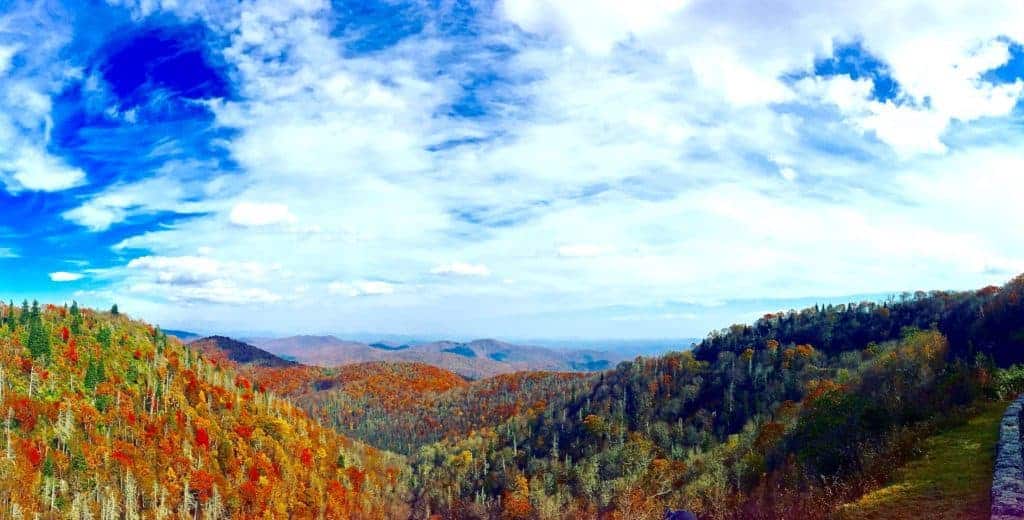 A panoramic view of rolling hills covered in autumn-colored trees surrounds cozy cabins in Asheville, NC under a bright blue sky with scattered white clouds, creating a scenic landscape.