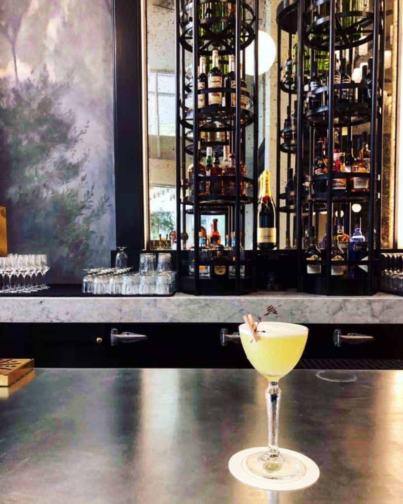 A yellow cocktail garnished with a sprig sits on a white coaster atop a shiny bar counter, set against the backdrop of Charlotte food culture, with a marble bar and tall shelves of liquor bottles in the background.