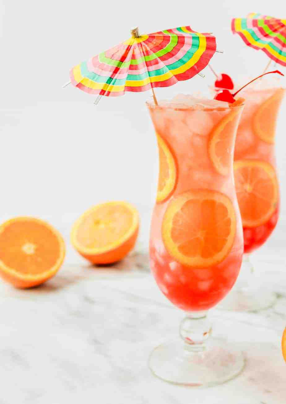 two hurricane drinks in hurricane glasses with orange slices, cherries and rainbow umbrellas 