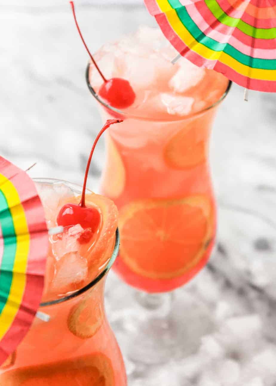 two hurricane drinks in hurricane glasses topped with cherries and a rainbow umbrella