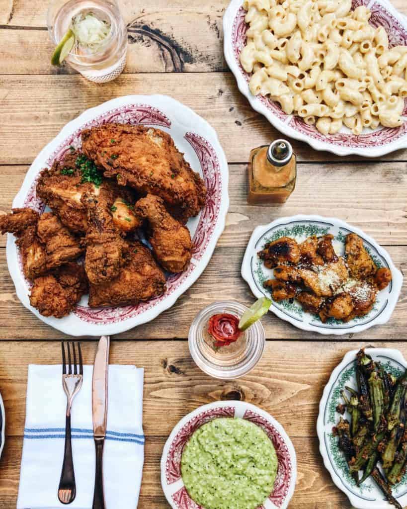 A wooden table set with plates of fried chicken, macaroni and cheese, roasted vegetables, green sauce, drinks with lime, and neatly arranged cutlery on a napkin—classic comfort fare found in top Charlotte restaurants.