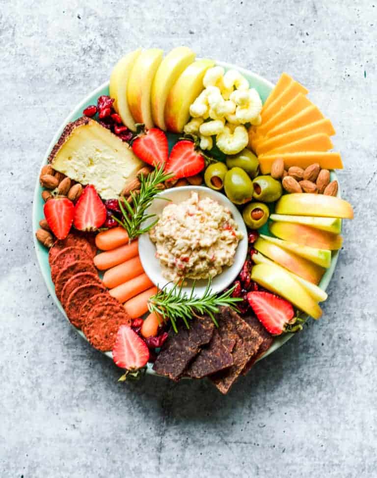 A simple cheese platter featuring sliced apples, cheese, olives, almonds, carrots, strawberries, crackers, and a bowl of dip in the center—all arranged neatly on a light gray surface.
