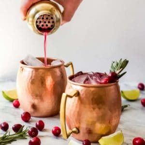 A hand pours red liquid from a cocktail shaker into a copper mug filled with ice, beside another mug garnished with rosemary and cranberries—an inviting scene for those exploring basic cocktail recipes. Lime wedges, cranberries, and rosemary sprigs rest on the marble surface.