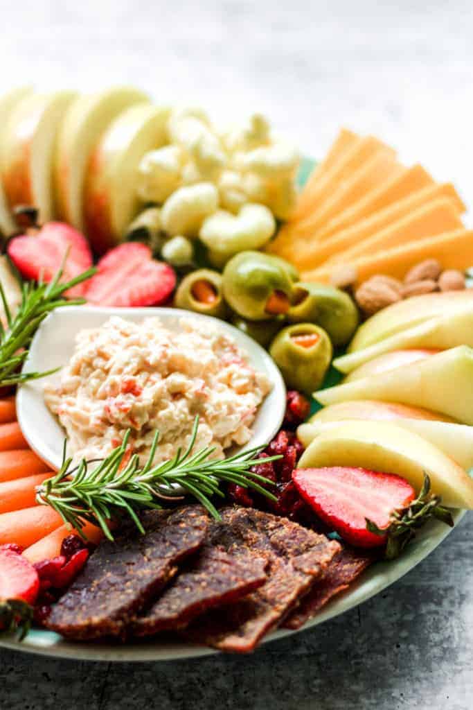 pimento cheese in a white bowl surrounded by beef jerky, cheddar cheese, and apple slices