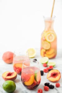 carafe of white wine sangria with a wooden spoon in it in the background with two glasses of sangria and fruit