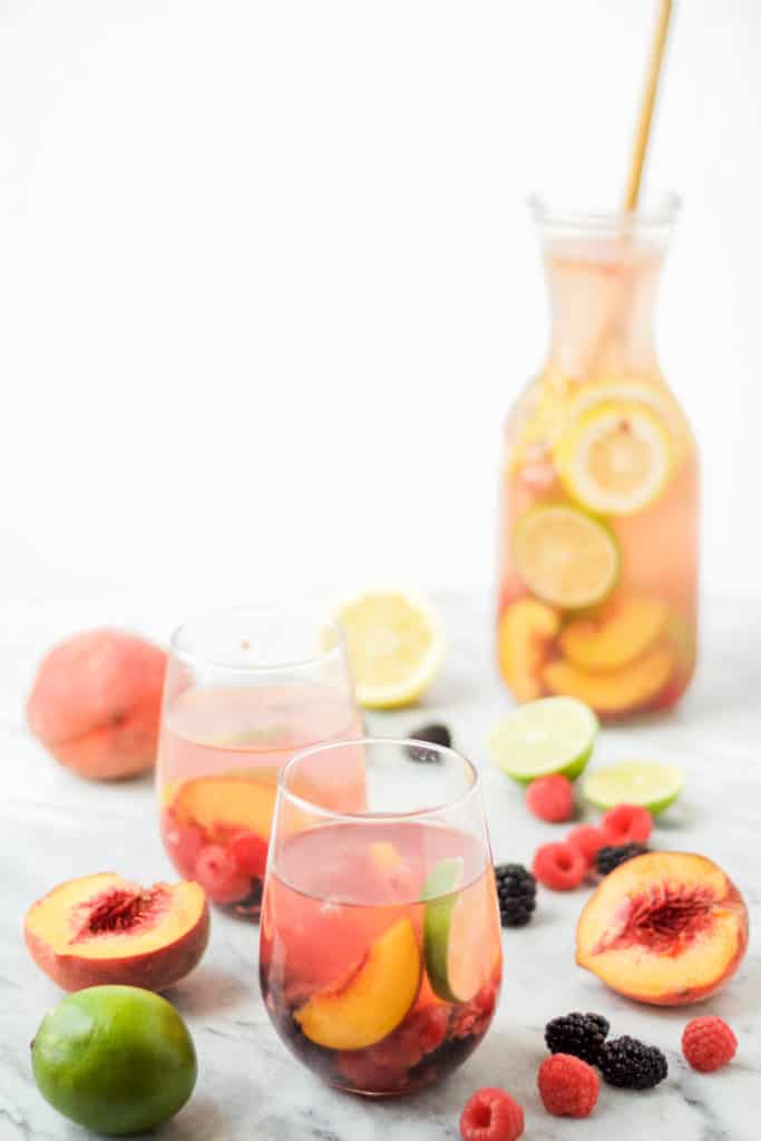 carafe of white wine sangria with a wooden spoon in it in the background with two glasses of sangria and fruit