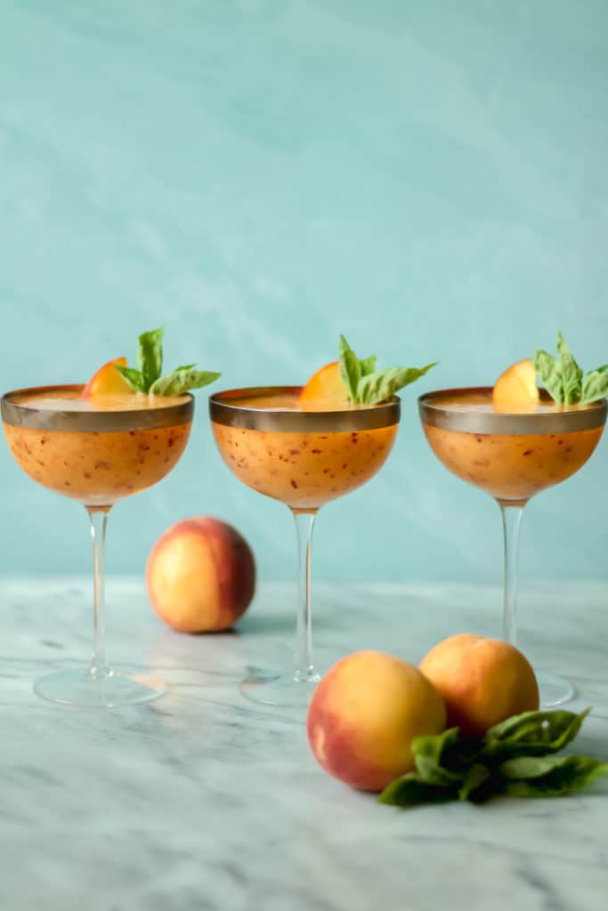 three cocktail glasses filled with peach frose with two peaches in front and one peach behind