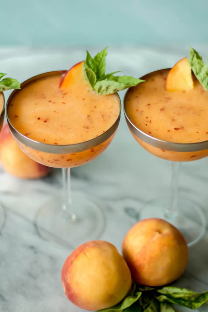 two cocktail glasses filled with peach frose garnished with green basil leaves against a blue background