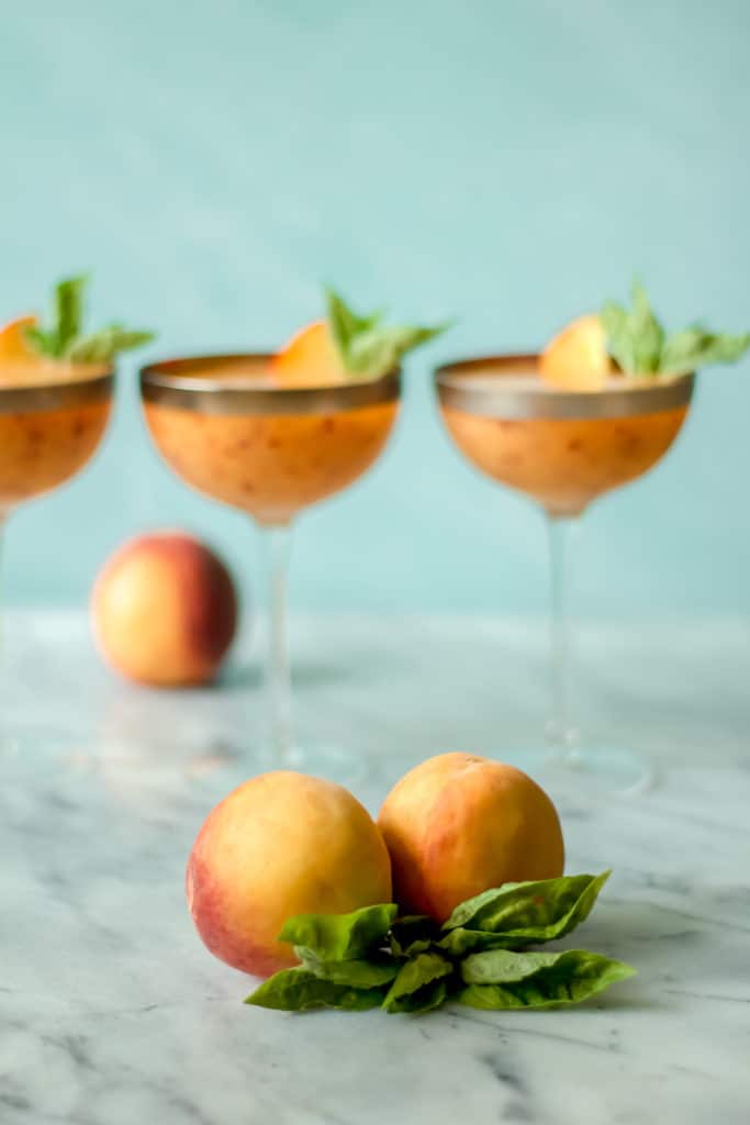 two peaches and a bunch of basil leaves with cocktails in the background