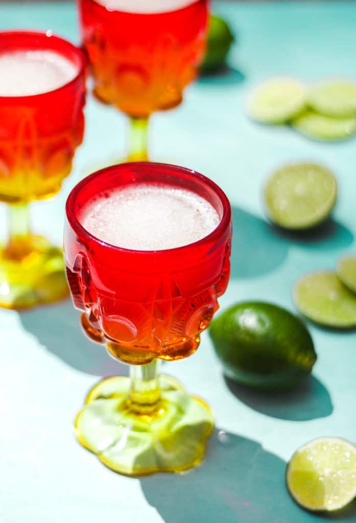 red and yellow cocktail glasses filled with frozen gin and tonic on a blue background with whole and sliced limes