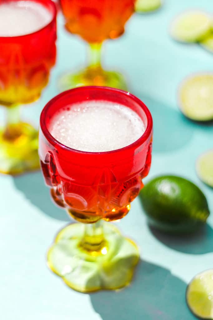 red and yellow stem glass with frozen drink on a blue background next to limes