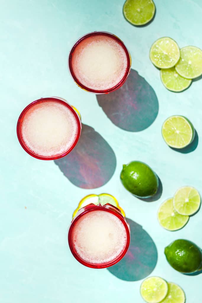 three frozen gin and tonic cocktails with white liquid in red glasses from overhead on a teal background with lime slices