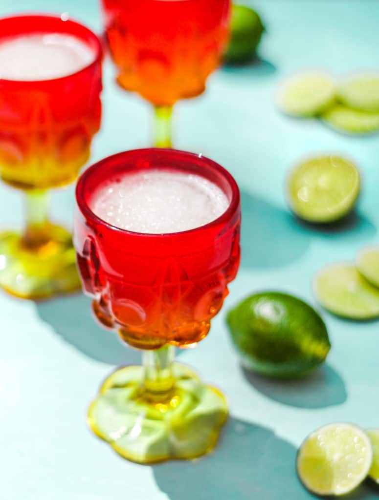 three red and yellow cocktail glasses filled with white slushie liquid on a blue background next to whole and sliced limes