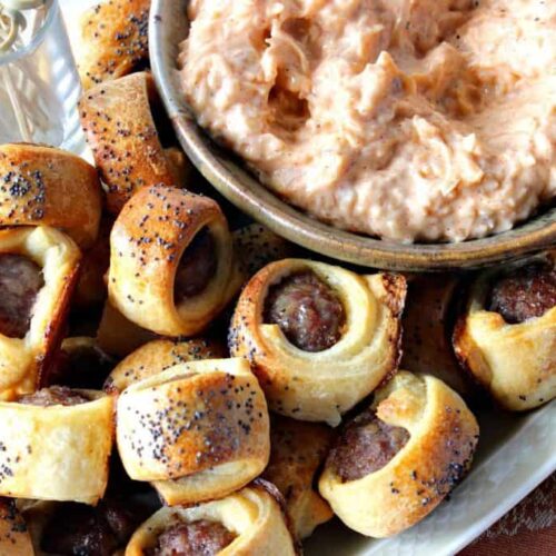 A plate of pigs in a blanket sprinkled with poppy seeds, served beside a bowl of creamy, orange-colored dipping sauce—one of the ultimate football food favorites.