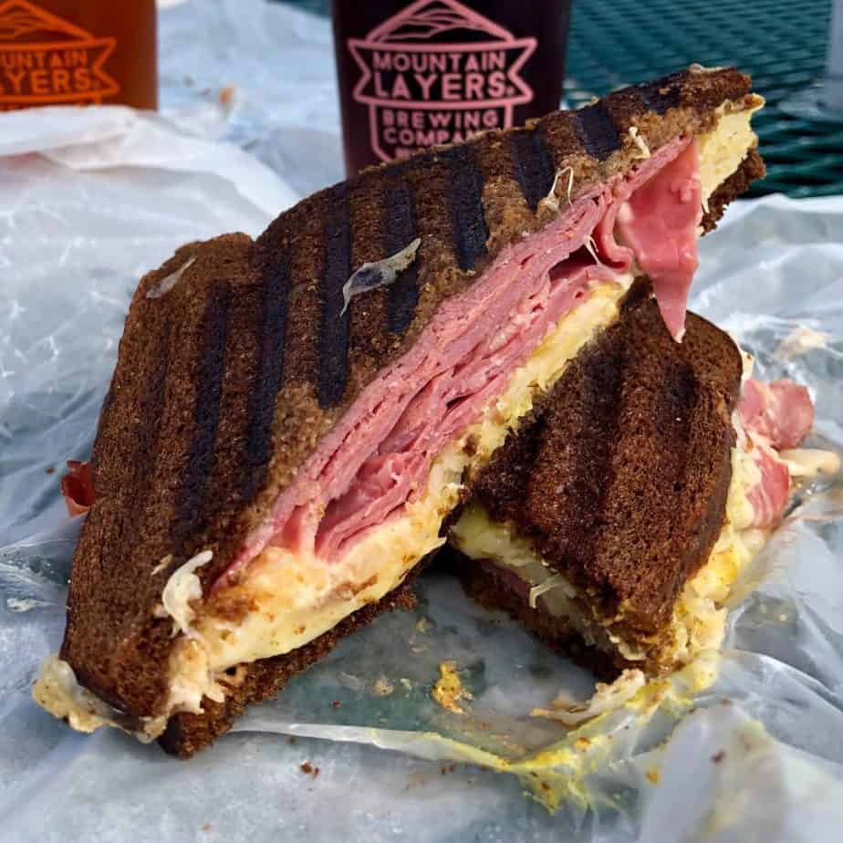 two halves of a ruben sandwich with pumpernickel bread stacked on top of each other with a beer glass and pink logo in the background