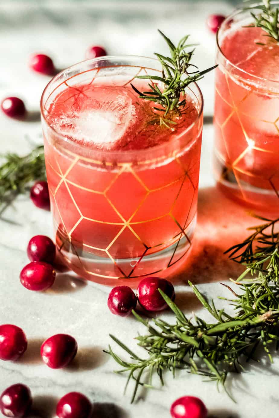 glass with gold designeds filled with pink colored cranberry bourbon smash with whole cranberries and rosemary on a marble background