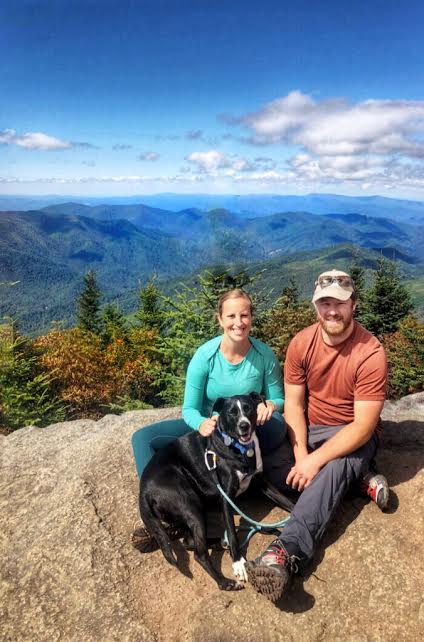 man in an orange shirt, woman in a green shirt and black and white dog in front of mountain views on a hike in north carolina