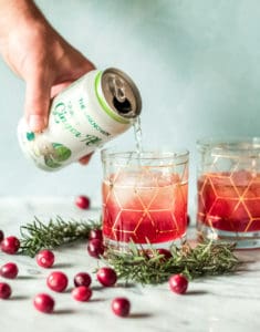 A hand pours ginger ale from a can into a glass with ice and red liquid for a cranberry bourbon smash recipe. Another glass, cranberries, and rosemary sprigs sit on the marble surface. Both glasses feature a gold geometric pattern.
