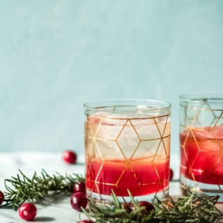 Two glasses with geometric gold patterns filled with a red and clear layered drink, inspired by a cranberry bourbon smash recipe, garnished with rosemary sprigs and surrounded by fresh cranberries on a light surface with a soft blue background.