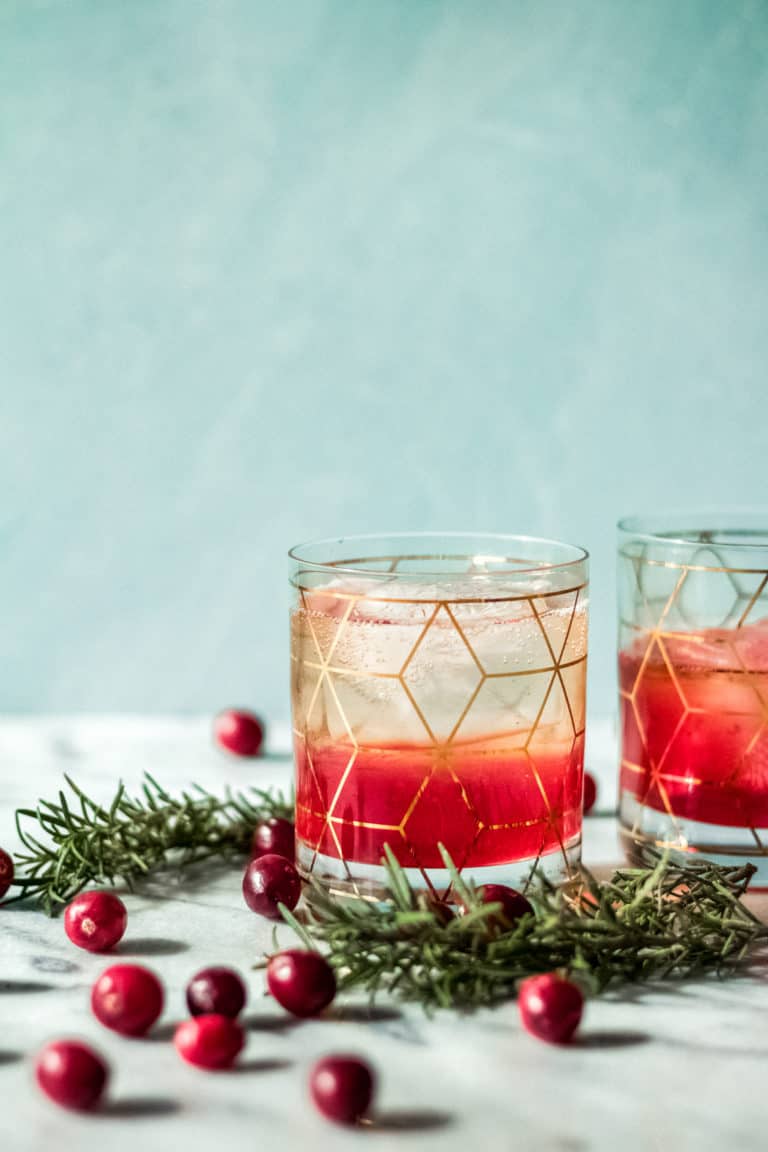 Two glasses with geometric gold patterns filled with a red and clear layered drink, inspired by a cranberry bourbon smash recipe, garnished with rosemary sprigs and surrounded by fresh cranberries on a light surface with a soft blue background.