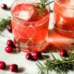 Two glasses of a pink cranberry bourbon smash recipe with ice, garnished with rosemary sprigs, sit on a marble surface surrounded by fresh cranberries and additional rosemary. The glasses feature a gold geometric pattern.