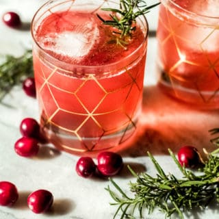 Two glasses of a pink cranberry bourbon smash recipe with ice, garnished with rosemary sprigs, sit on a marble surface surrounded by fresh cranberries and additional rosemary. The glasses feature a gold geometric pattern.
