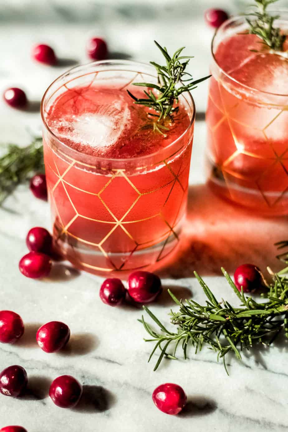 two glasses with gold designeds filled with pink colored cranberry bourbon smash with whole cranberries and rosemary on a marble background