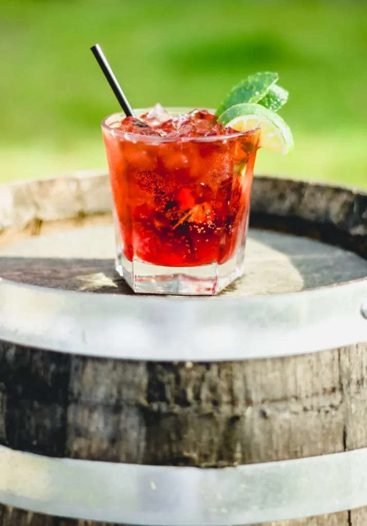 red cocktail with mint and lime wedge garnish on a whiskey barrel with a green backgroun red cocktail with mint and lime wedge garnish on a whiskey barrel with a green backgroun