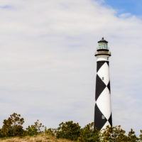A tall lighthouse with a black and white diamond pattern stands on a sandy, grassy hill with small trees under a partly cloudy sky, capturing the charm often found at beaches near Charlotte NC.