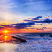 A vibrant sunset over the ocean, with the sun low on the horizon and waves crashing onto the shore, evokes scenes from beaches near Charlotte NC, as the sand reflects bright orange, pink, and blue hues beneath colorful clouds.