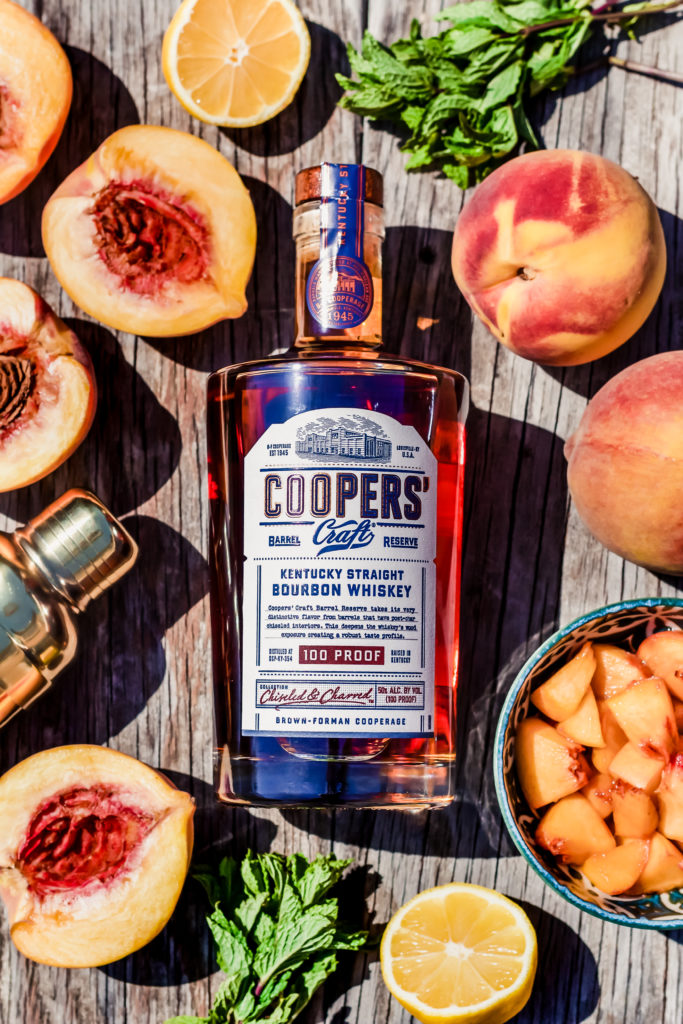 bottle of coopers craft barrel reserve bourbon on a wooden table surrounded by peaches slices in half mint lemon and a cocktail shaker