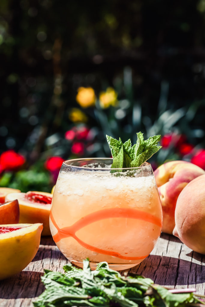 glass with peach cocktail filled with crushed ice on a table surrounded by peaches