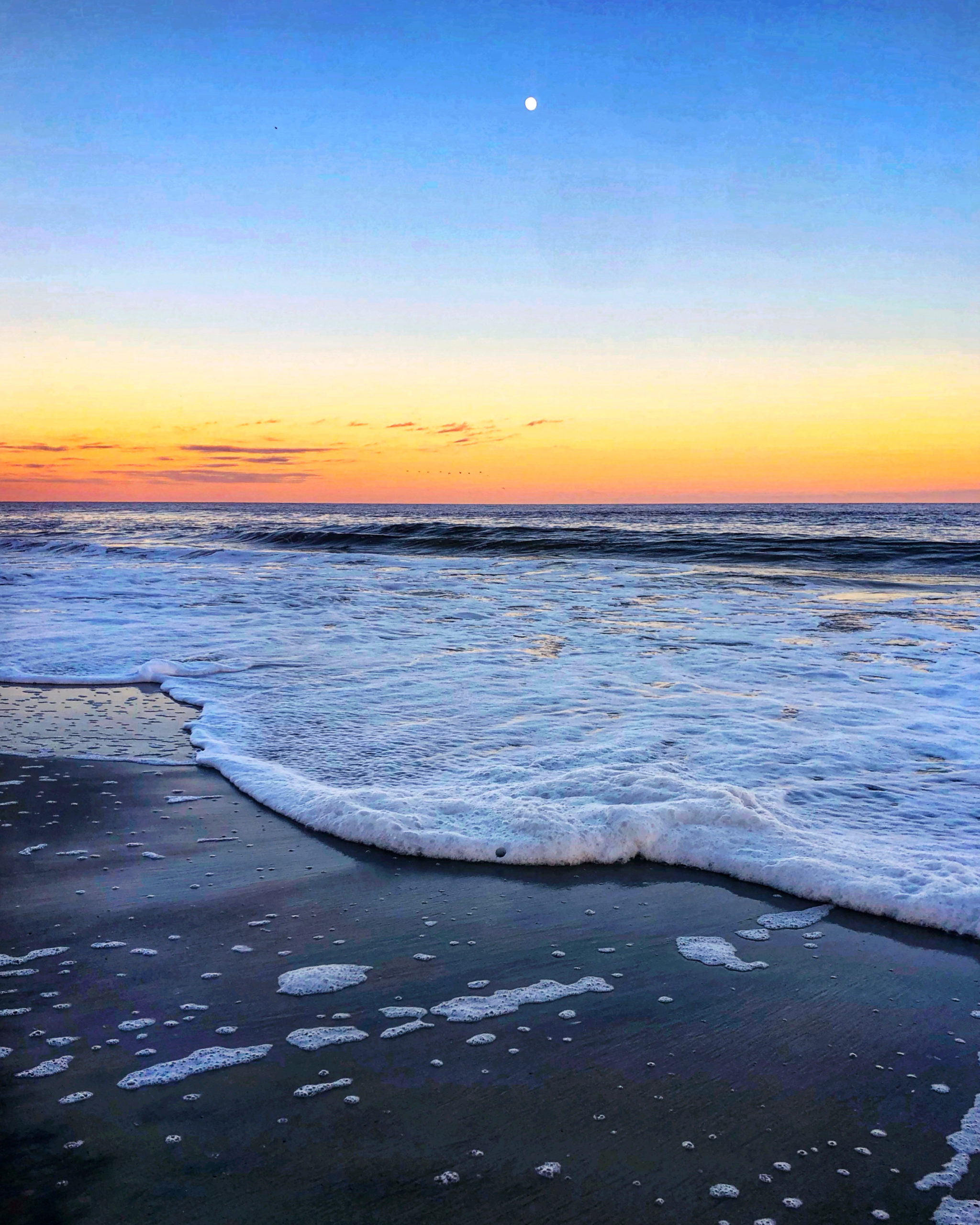 Waves gently wash onto a sandy beach at sunset, with the sky glowing in shades of orange and blue. The moon is visible above the horizon, and foam from the waves dots the beach. 