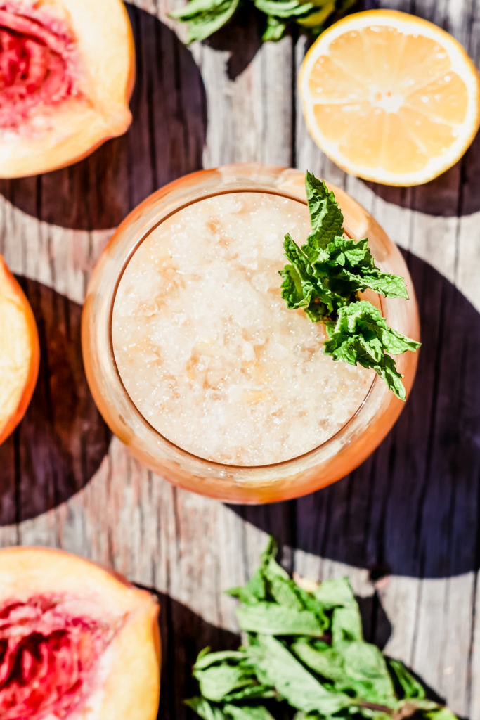 overhead picture of a cocktail glass with crushed ice and a mint garnish surrounded by peach halves, mint and lemon halves