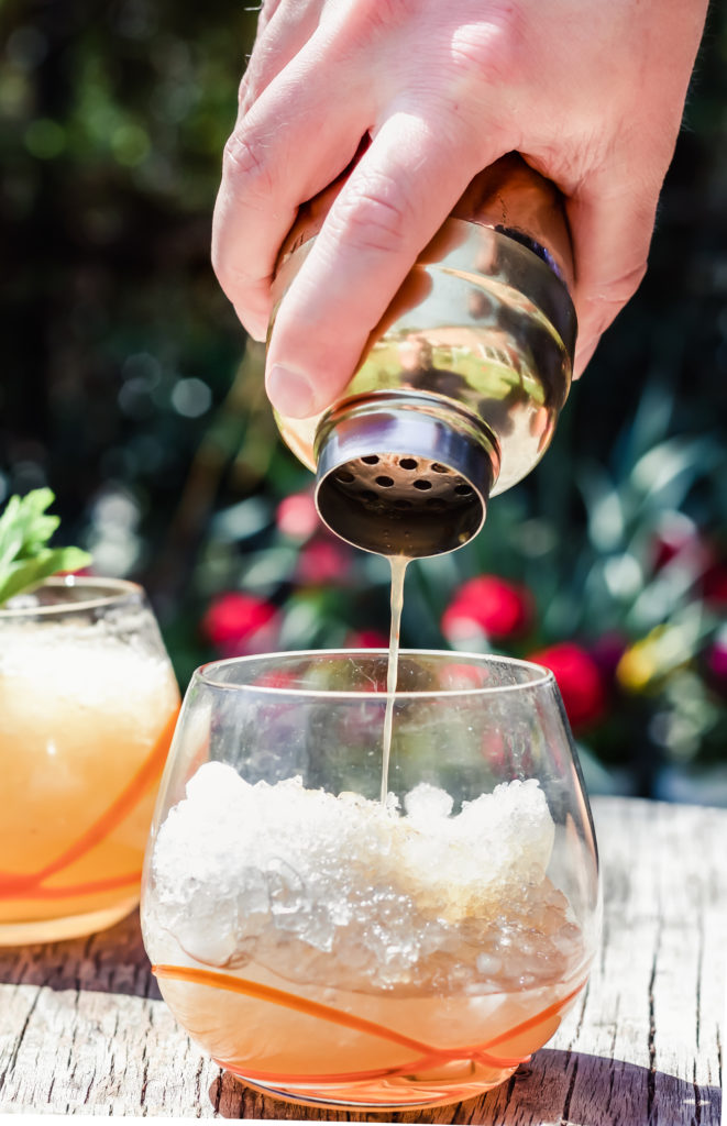 pouring peach whiskey smash recipe into a cocktail glass filled with ice from a gold cocktail shaker