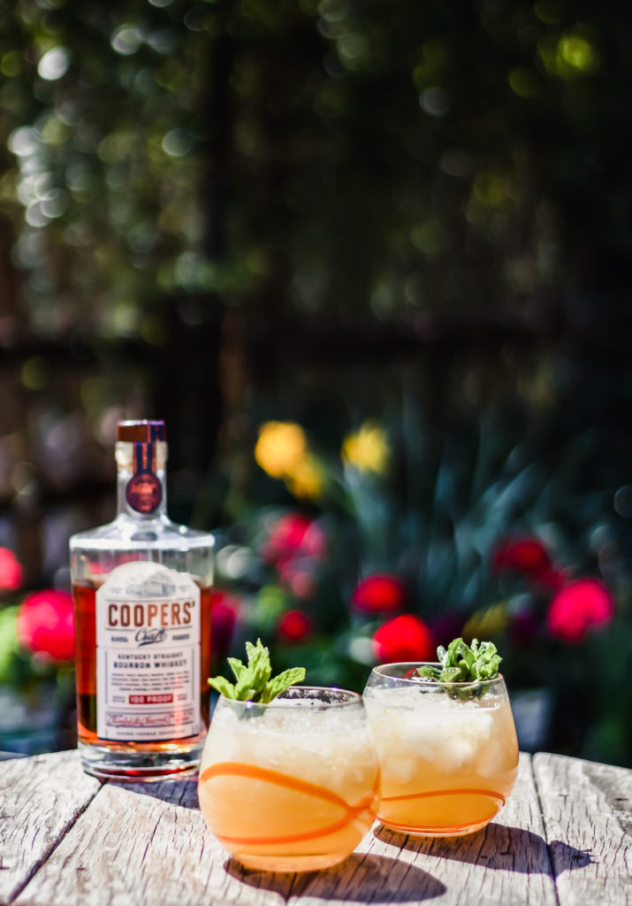 two bourbon peach smash cocktails on a table next to a bottle of coopers craft bourbon with flowers in the background