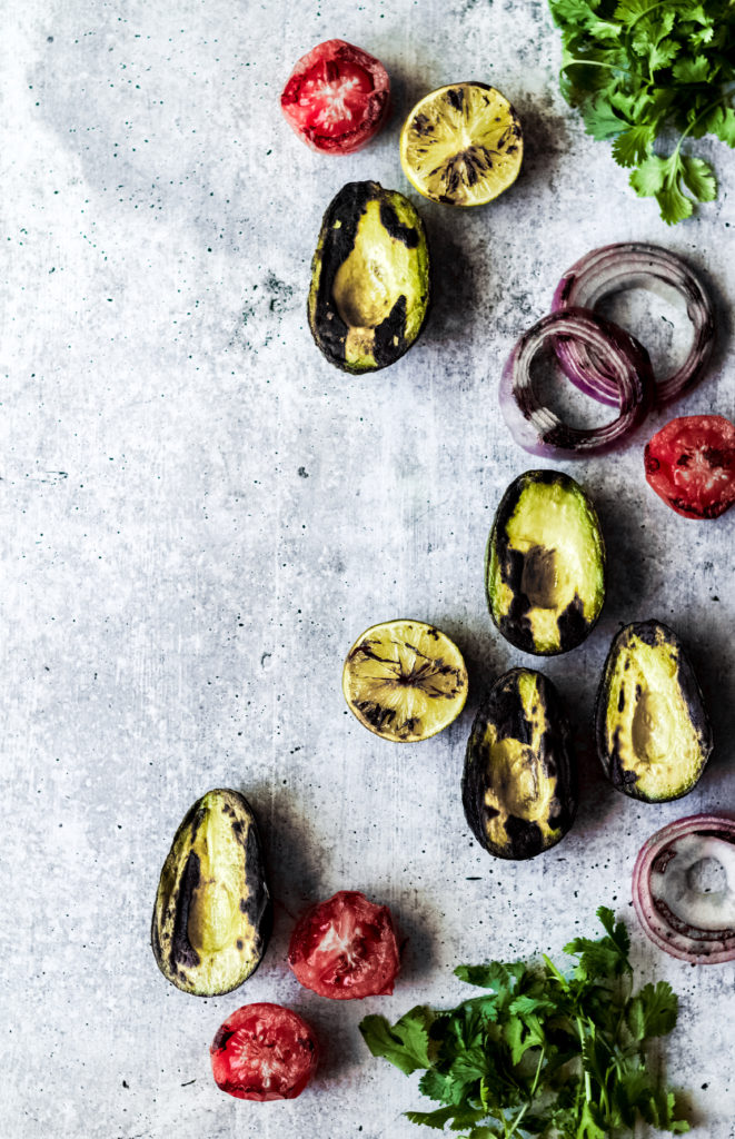 grilled veggies to make vegan guacamole including avocado halves, tomatoes, red onions and cilantro on a gray background