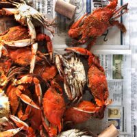 Steamed crabs covered in seasoning are spread out on newspaper with wooden mallets nearby, ready for eating. Shrimp and other seafood are also visible around the edges.