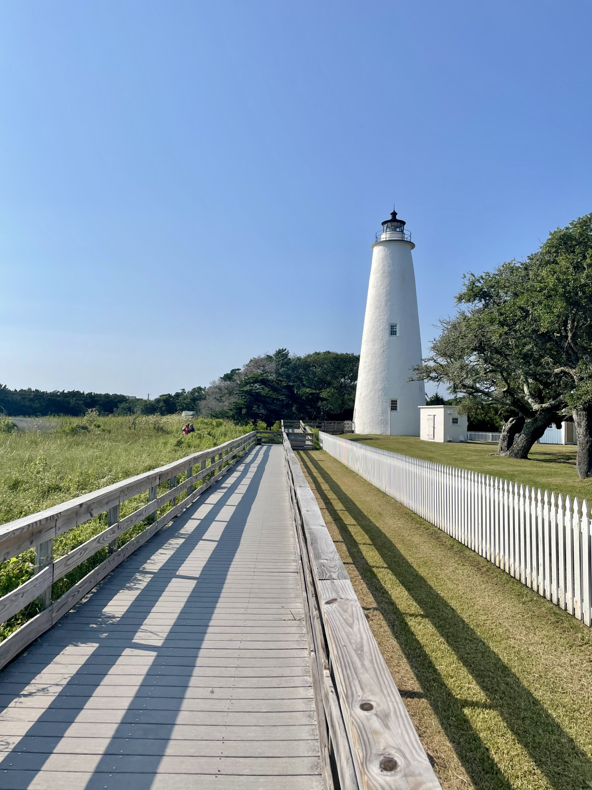 A wooden boardwalk leads to a tall white lighthouse, bordered by a white picket fence and grassy areas under a clear blue sky. Trees and greenery are visible in the background.