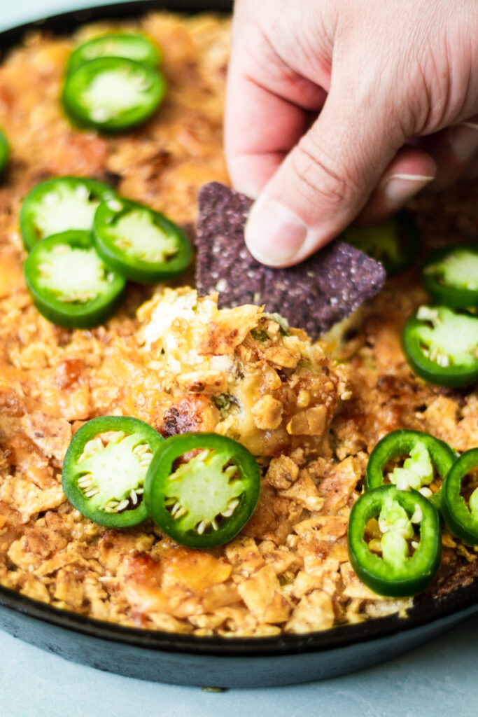 dipping a blue corn tortilla chip into a batch of jalapeno cream cheese dip with fresh jalapenos on top
