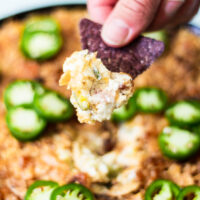 A hand holds a blue corn tortilla chip topped with cheesy dip and jalapeño slices above a platter of dip garnished with more fresh jalapeño slices.