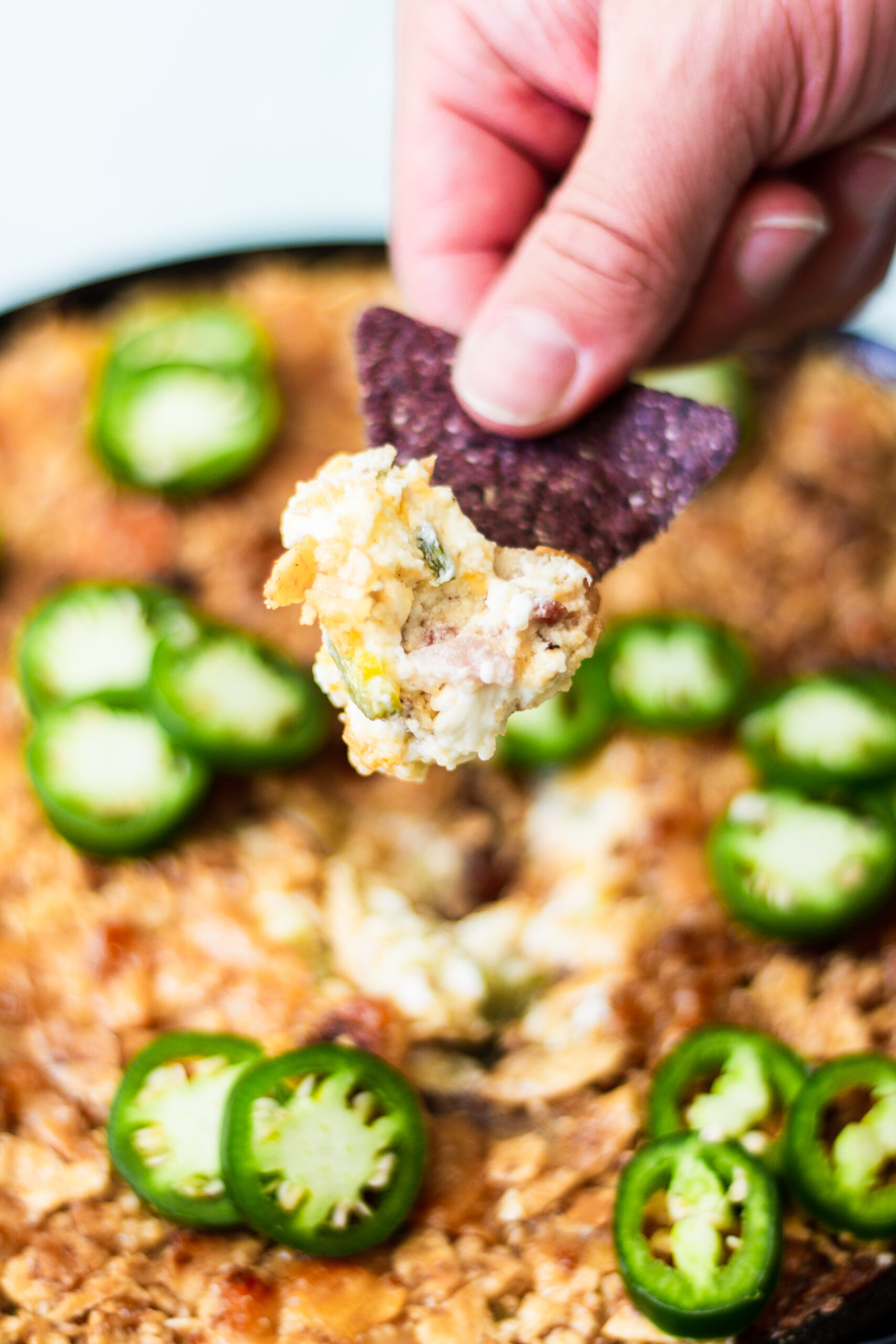 A hand holds a blue corn tortilla chip topped with cheesy dip and jalapeño slices above a platter of dip garnished with more fresh jalapeño slices.
