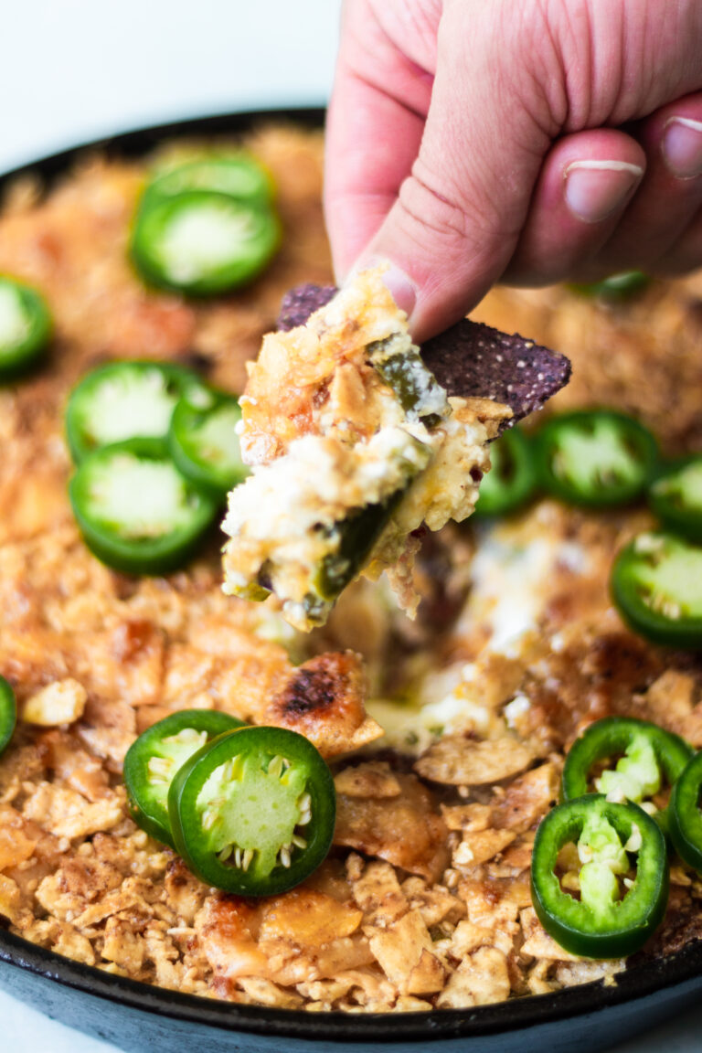 A hand dips a blue corn tortilla chip into a skillet brimming with creamy jalapeño popper dip, topped with fresh jalapeños—an irresistible twist on classic jalapeño cream cheese dip.
