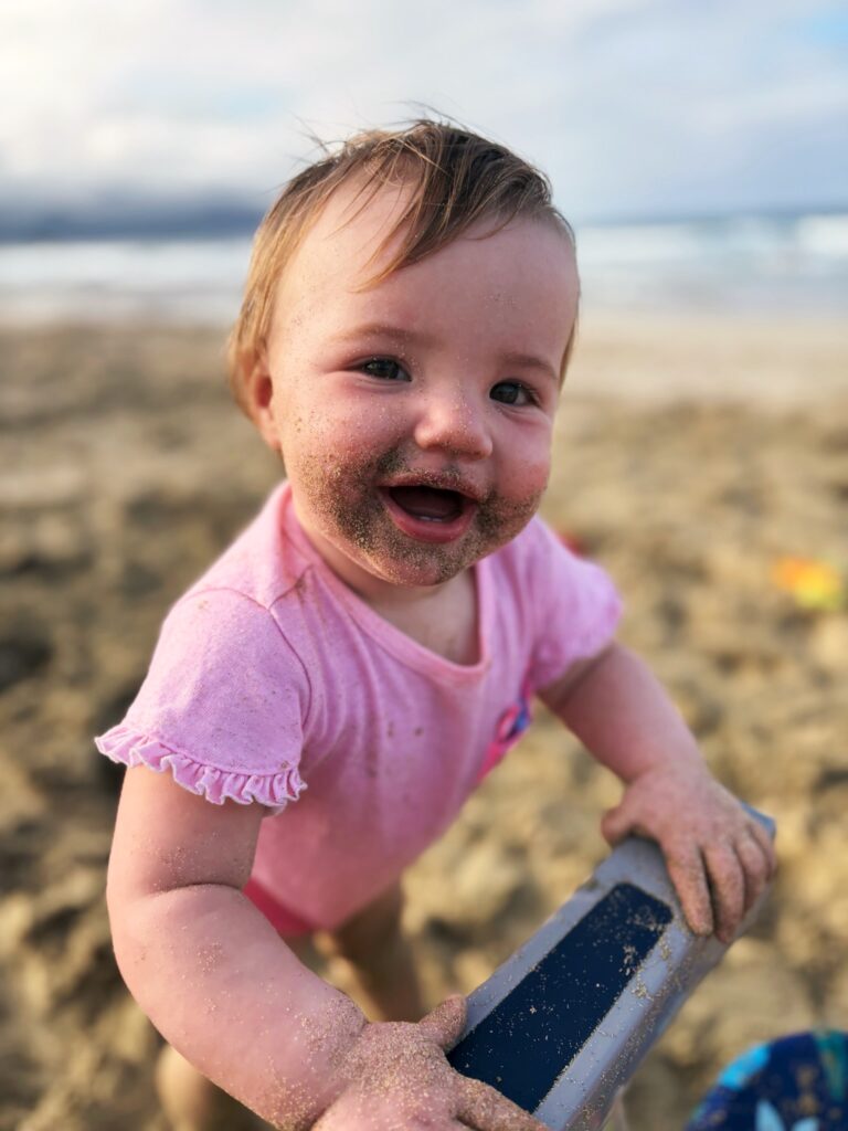 A smiling baby wearing a pink shirt stands on a sandy beach, with sand on their face and hands, holding onto a blue object, with the ocean and sky blurred in the background.