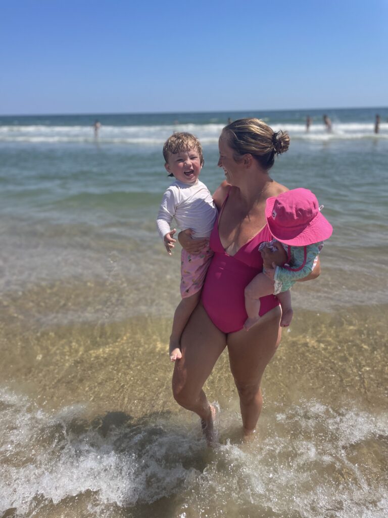 A woman in a pink swimsuit stands in shallow ocean water, smiling and holding a laughing toddler and a baby wearing a pink sunhat. The sky is clear and people are visible in the background near the waves.