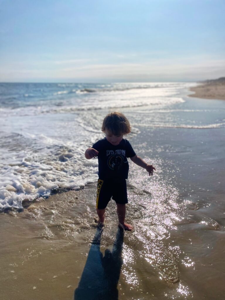 A young child in dark shorts and a graphic t-shirt walks along the wet sand at the beach, with gentle waves and sunlight reflecting on the water in the background.